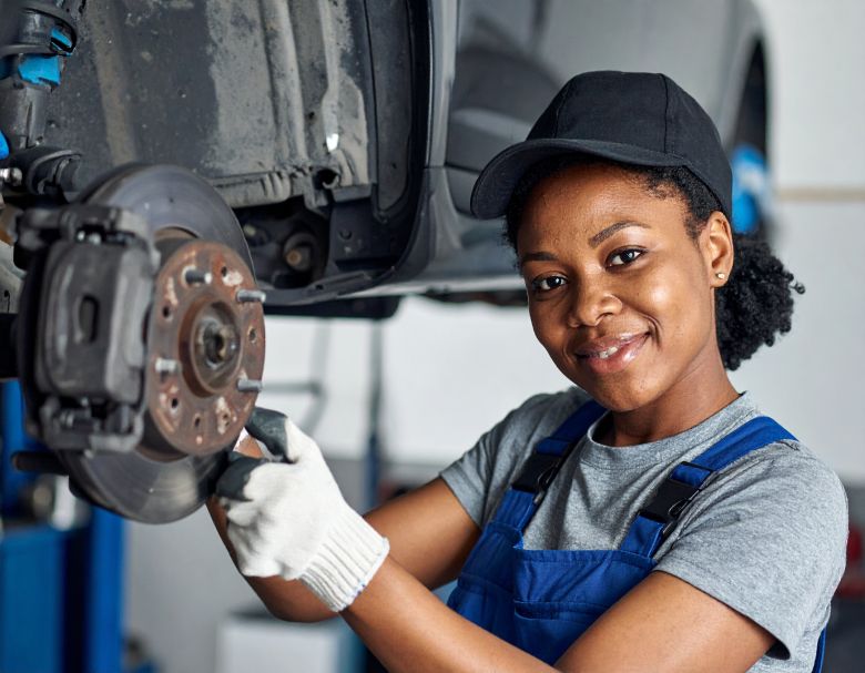 Mechanic working on car brakes; wearing gloves, hat, and overalls, smiling in a shop.