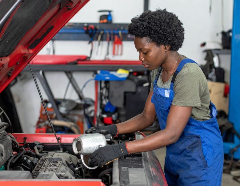 Woman in blue overalls working on a car engine in a garage.