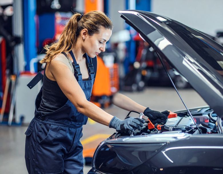 Woman in overalls working on a car engine in a garage; uses jumper cables.