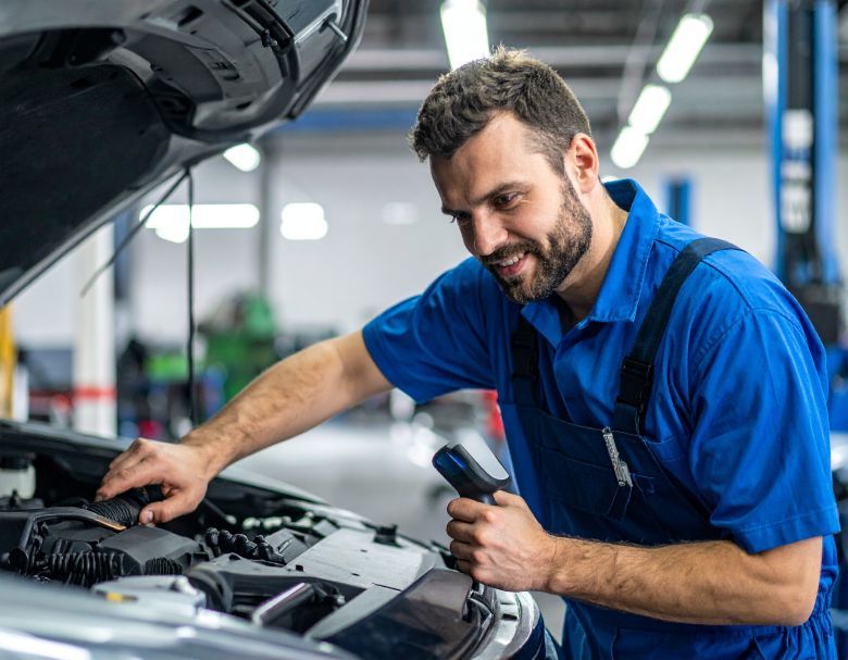 Mechanic in blue jumpsuit, smiling, inspecting car engine, holding a tool in an auto shop.