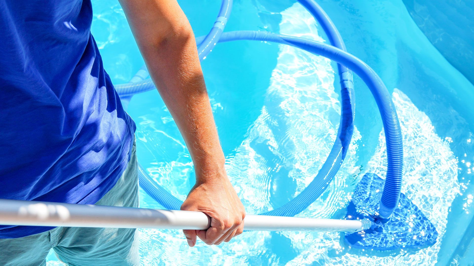 Person in blue shirt cleaning a blue swimming pool with a net and hose.