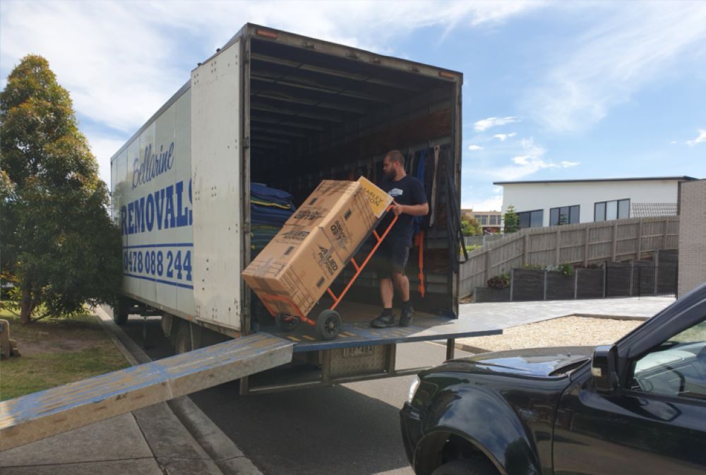 A man is pushing a large box on a dolly out of a moving truck.