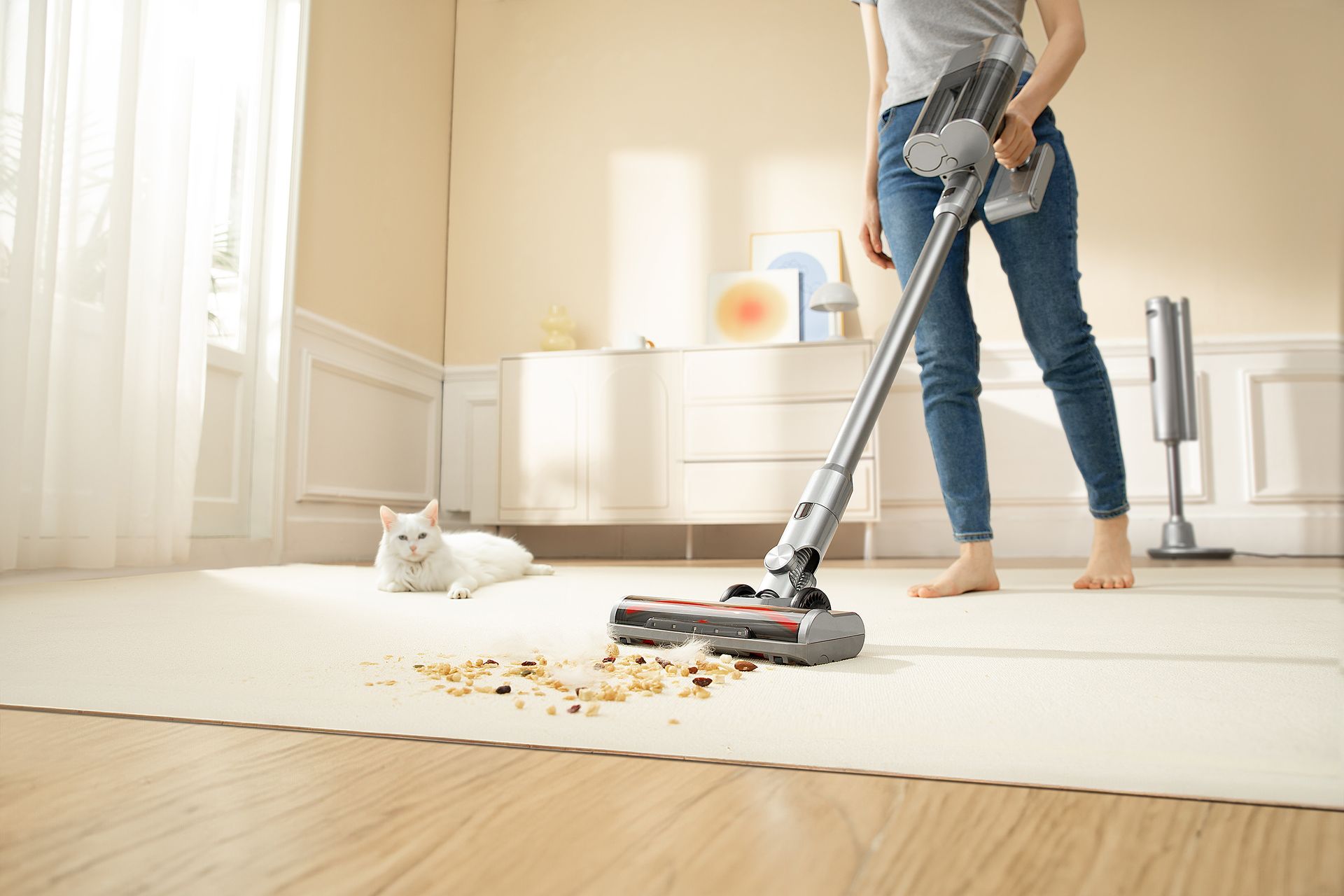 Woman vacuuming spilled food off a white carpet while a white cat watches nearby