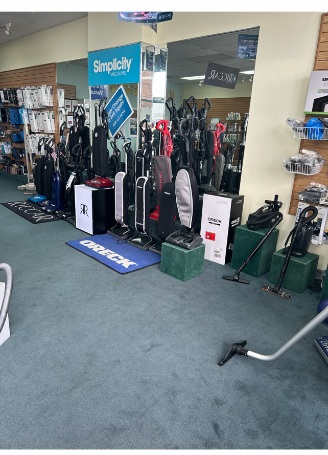 Vacuum cleaners displayed in a store, including upright and canister models. Blue carpet, green boxes, and a mirrored wall.