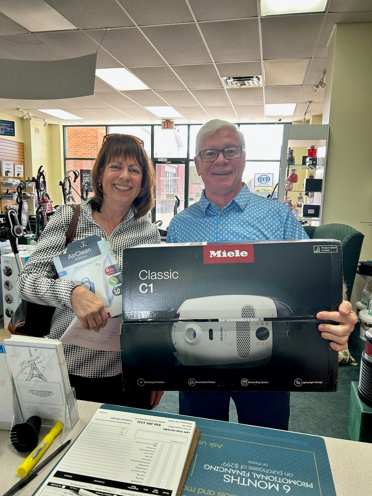 Couple in store holding a Miele Classic C1 vacuum cleaner box, smiling.