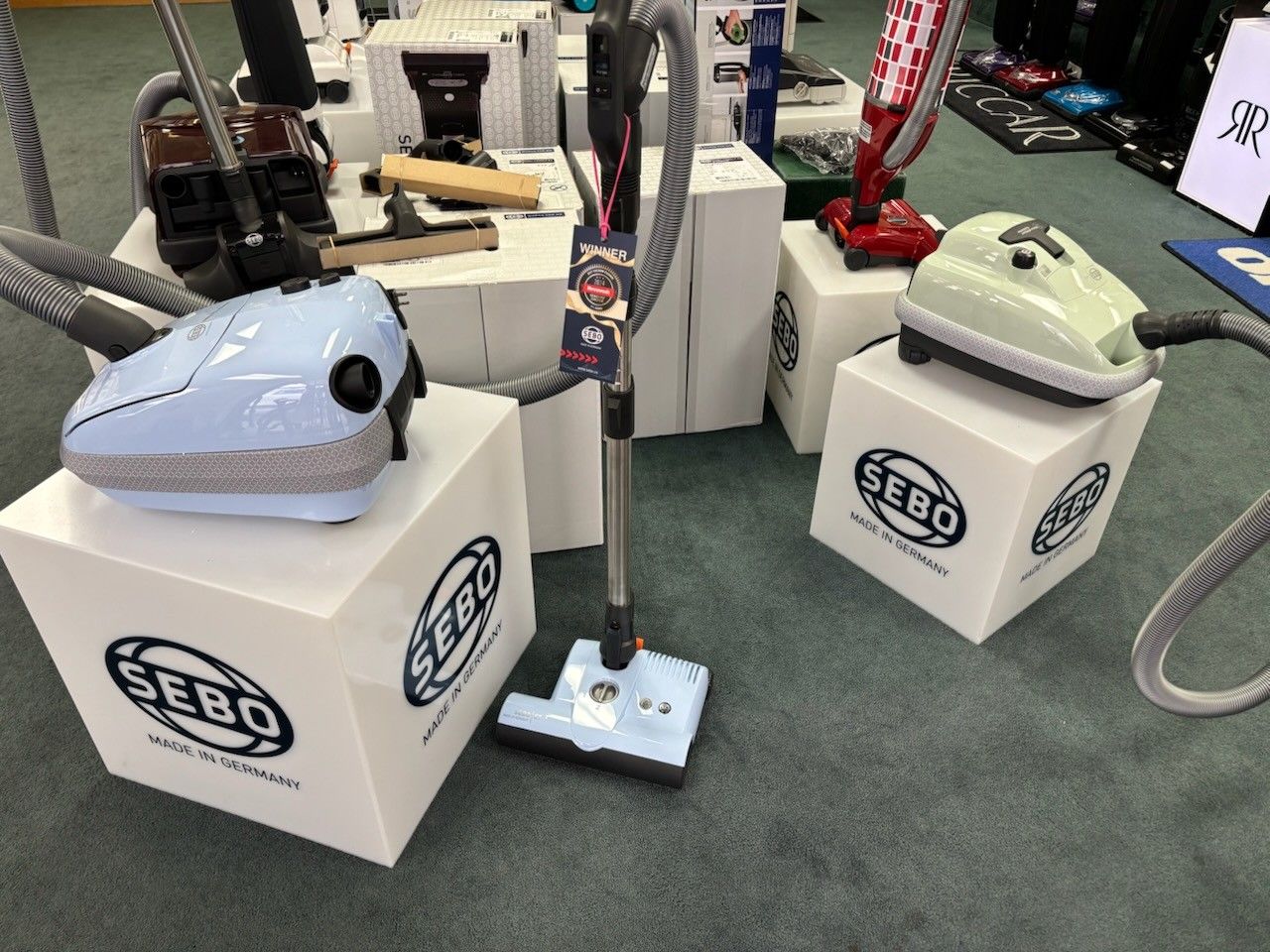 Display of SEBO vacuum cleaners on white cubes. Light blue, green, and red vacuums are featured.