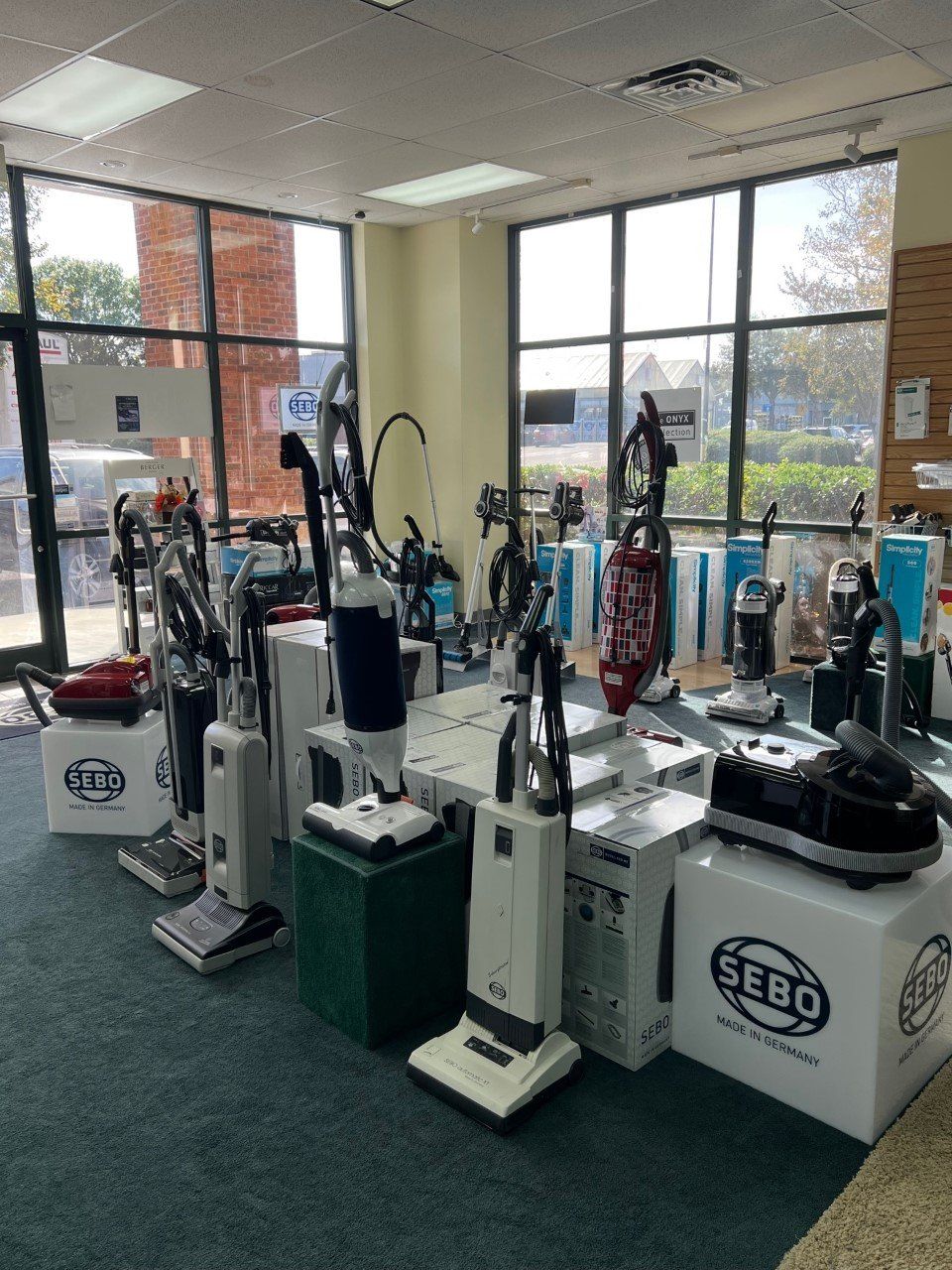 Vacuum cleaners displayed for sale inside a store with large windows and a green carpet.