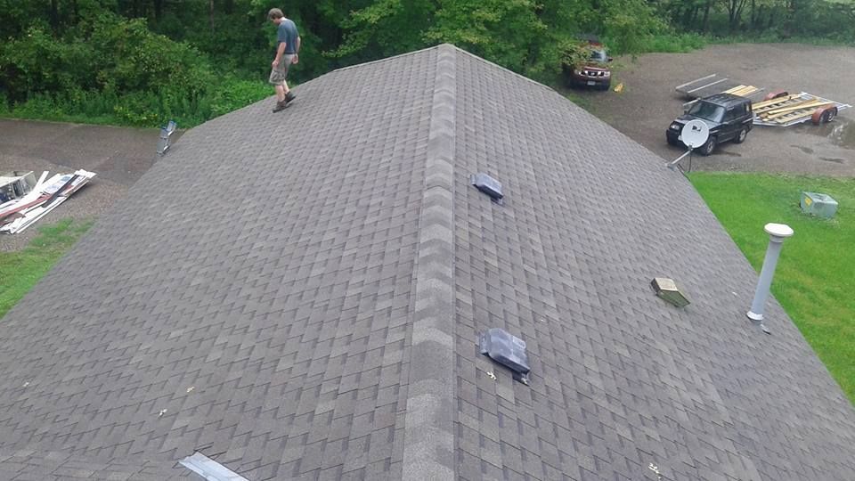 Roofing work on a shingled house roof, with a worker and materials visible on top and nearby grass and equipment below