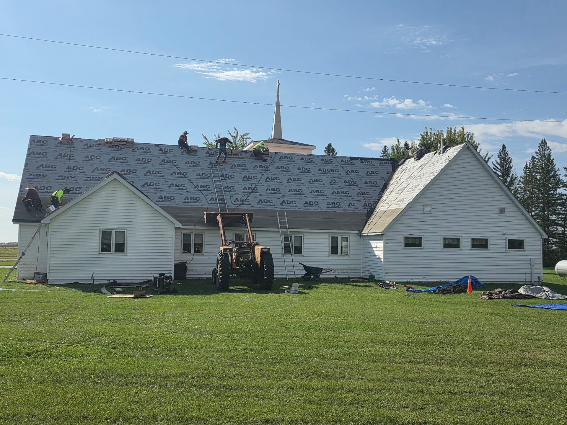 White house with damaged roof in a grassy field, with construction equipment in front under a blue sky