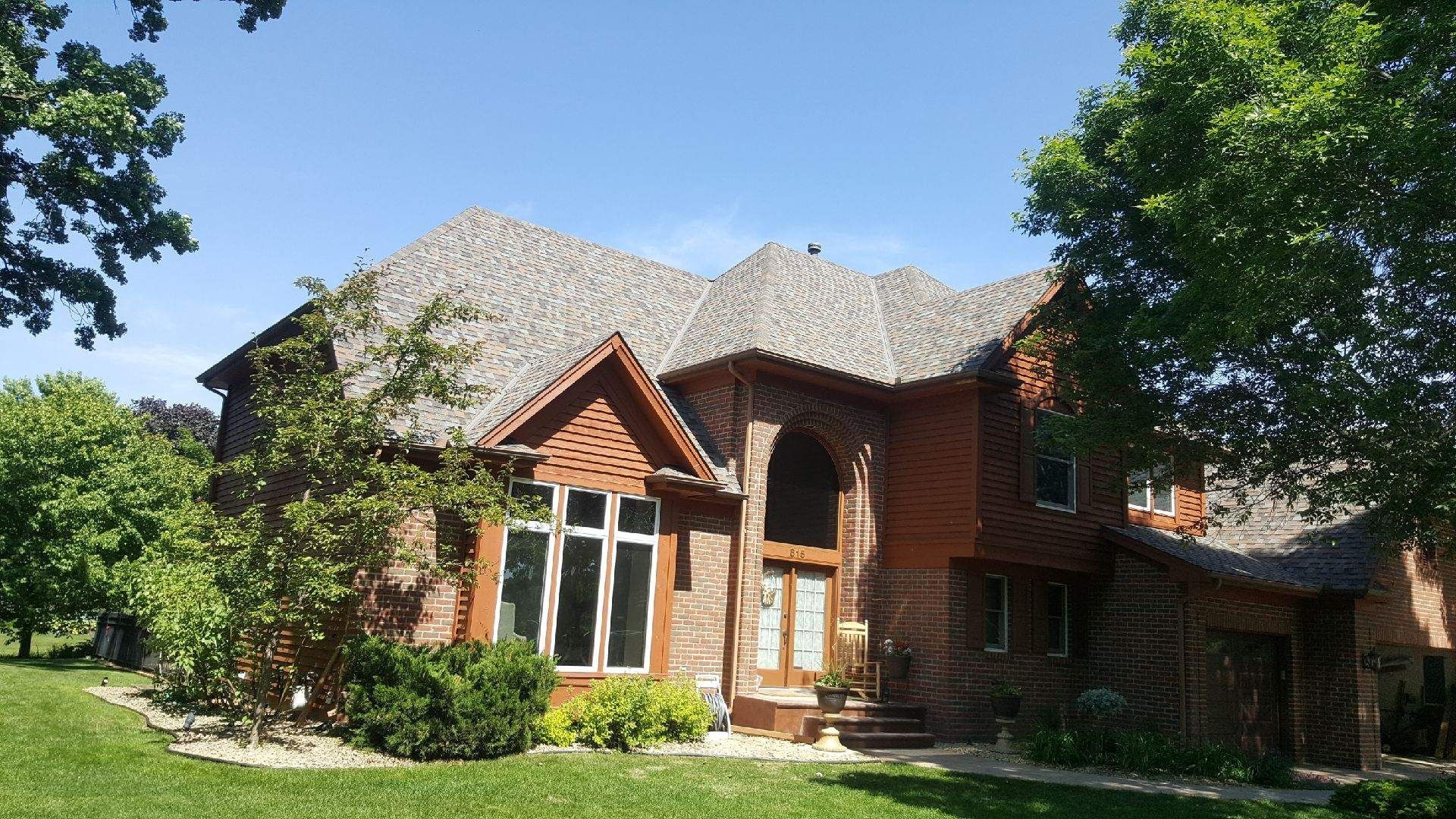 Brick house with a steep shingled roof, large windows, and a green lawn under bright sunlight