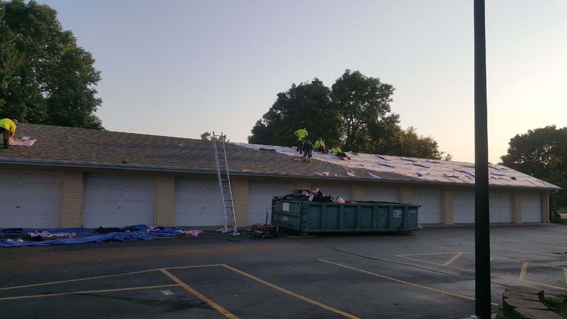 Empty parking lot beside a low beige building with a ladder and large dumpster at dusk