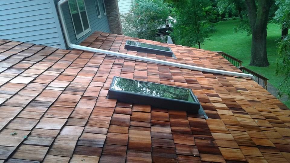 Roof covered in wet red-brown shingles with two skylights beside a house and green yard nearby