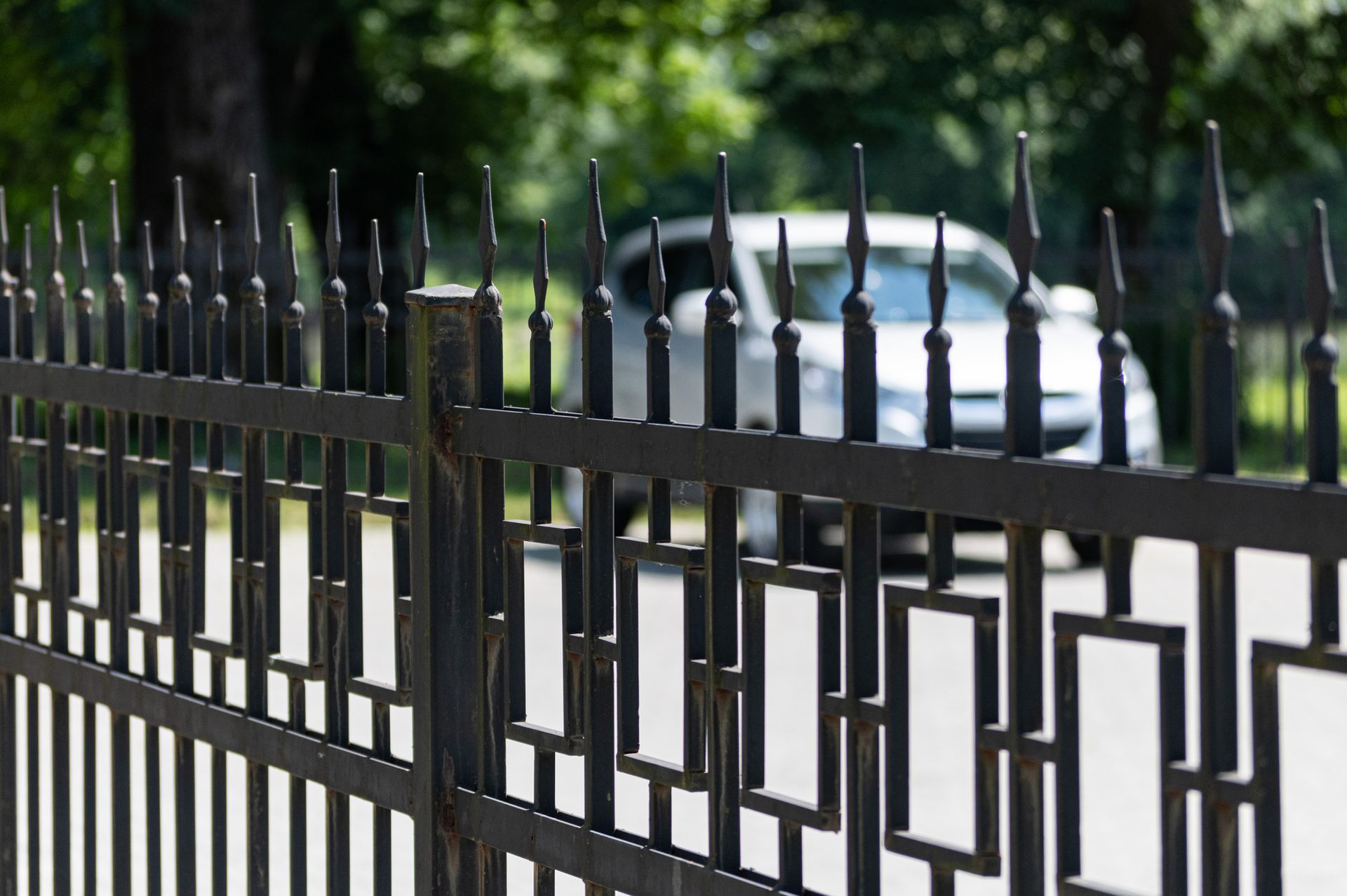 Black metal fence in foreground with a blurred white car behind it on a tree-lined street