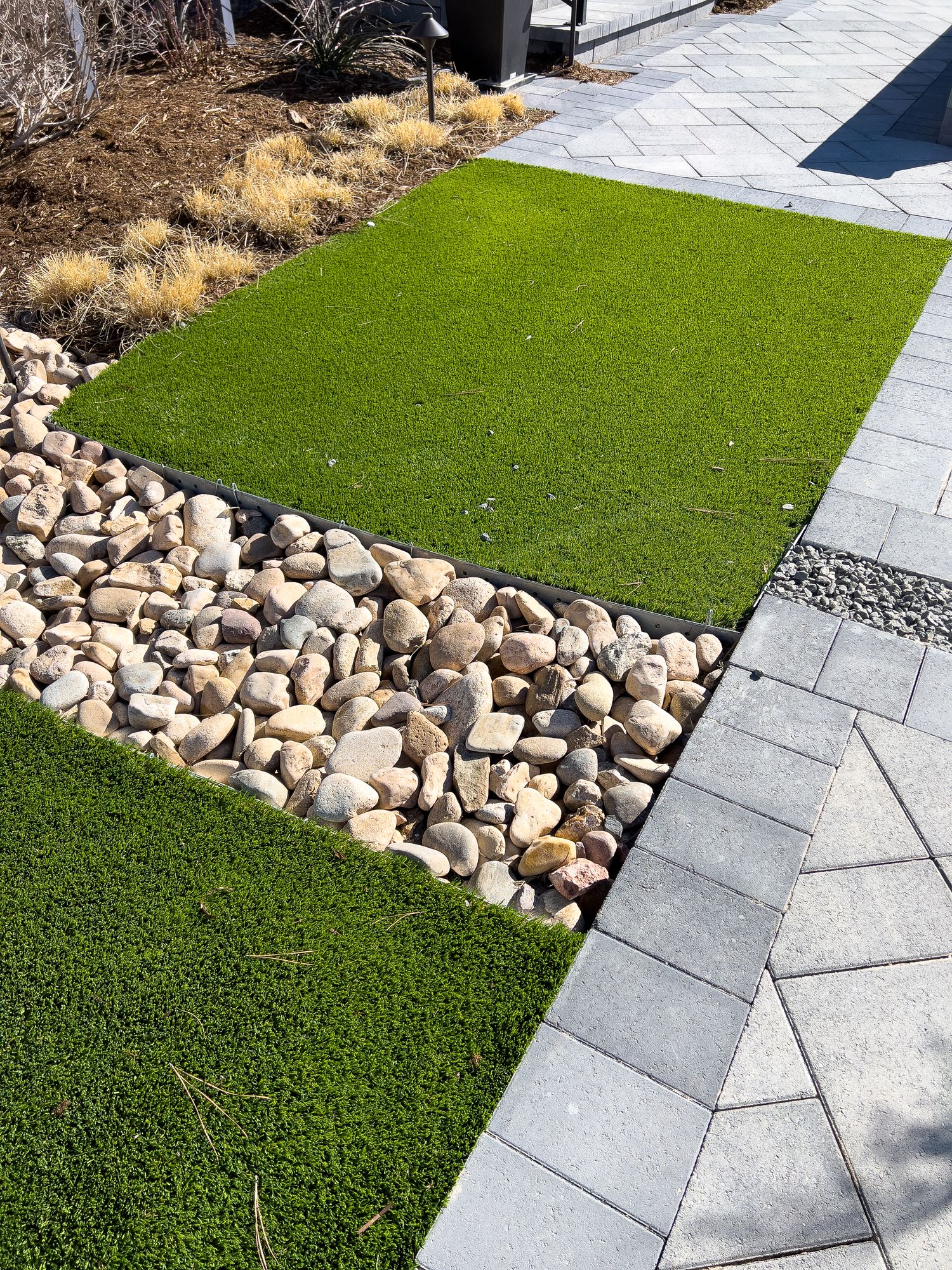 Modern backyard walkway with green turf panels, decorative rocks, and gray paver stones.