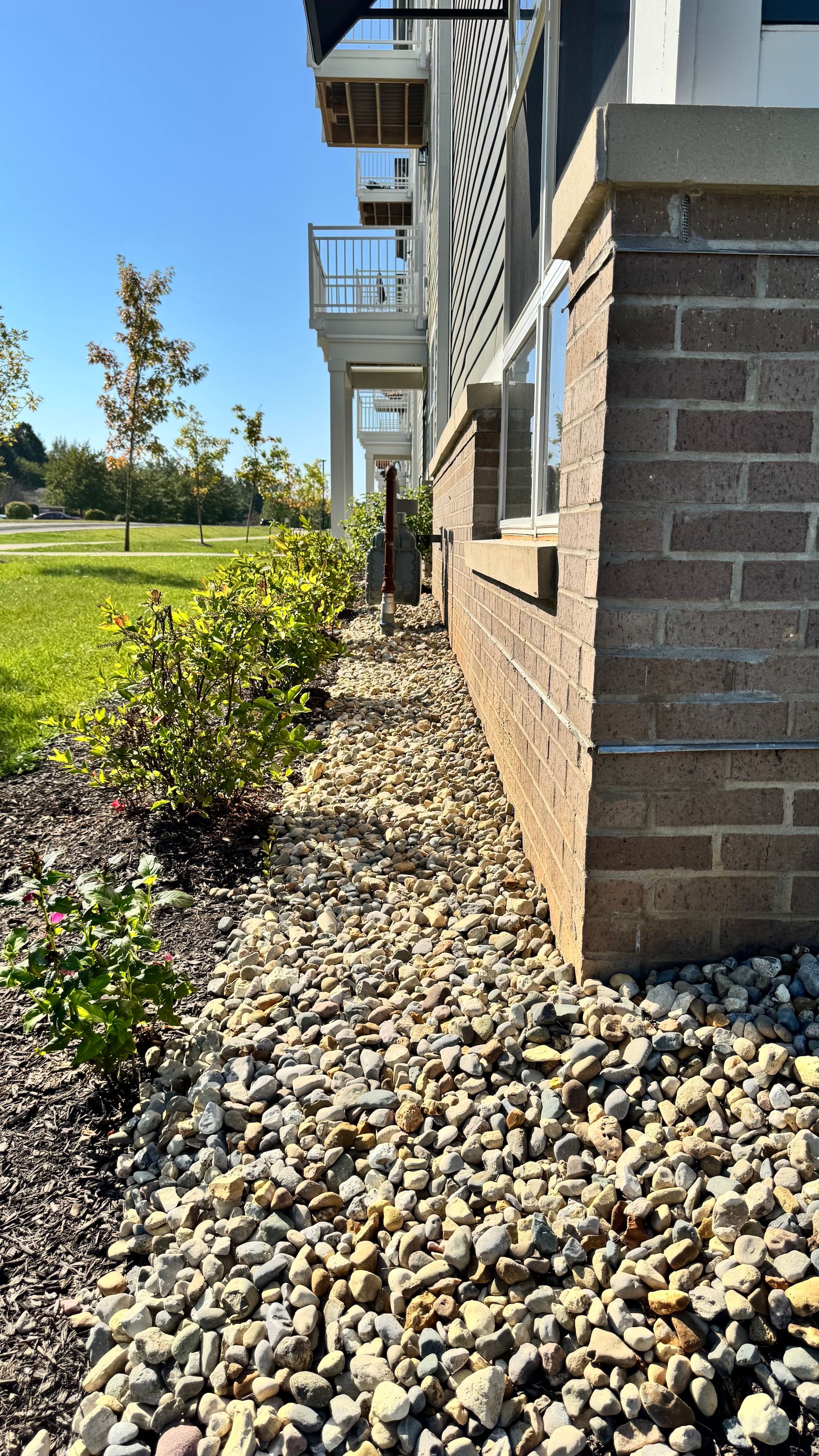 Side of an apartment building with rock landscaping, small shrubs, and a grassy lawn under a clear blue sky