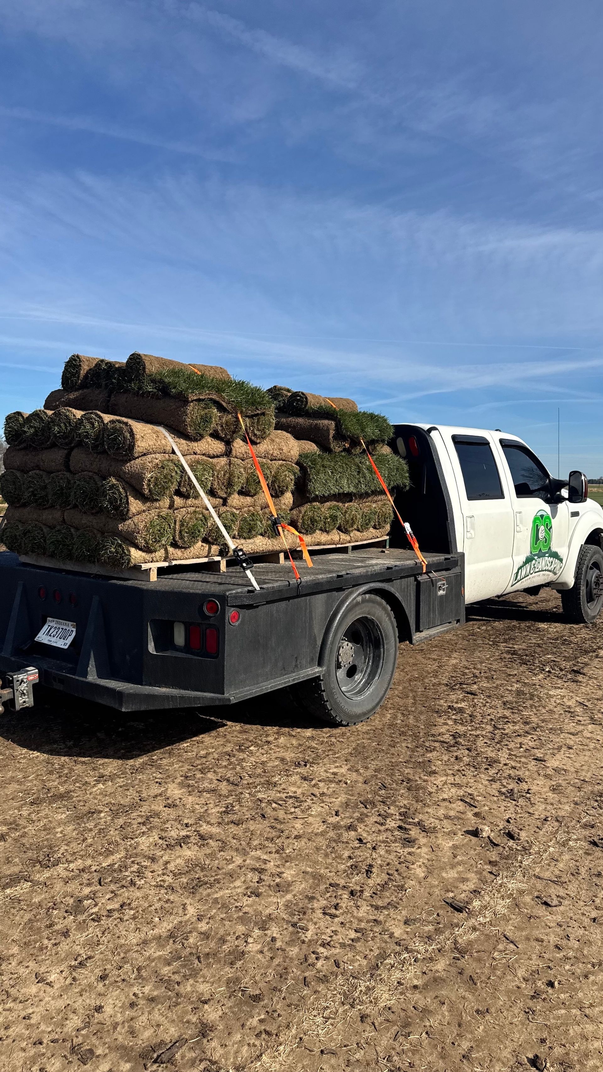 White flatbed truck loaded with stacked wood pallets in a dirt field under a blue sky