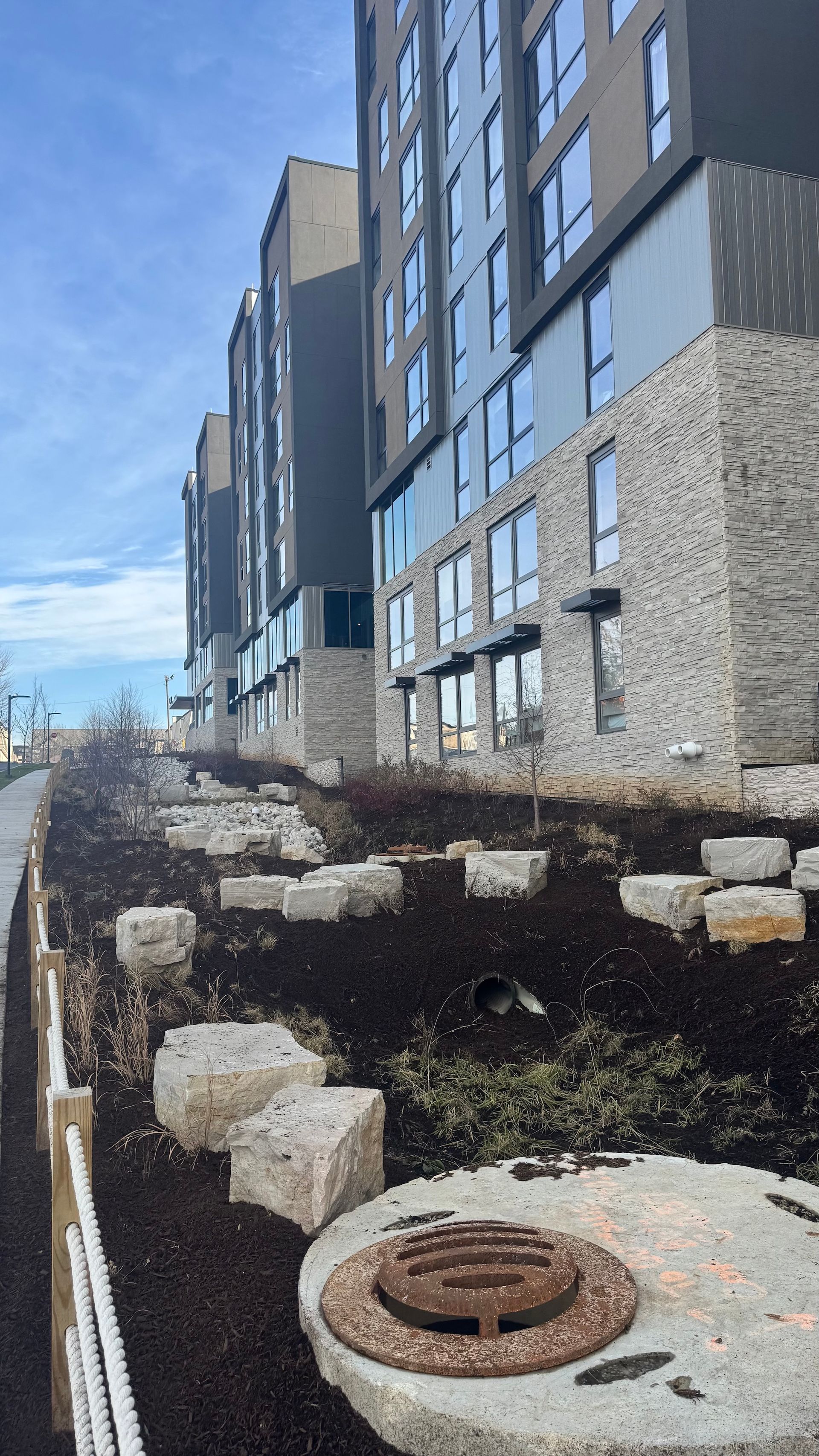 Apartment building beside a steep stone retaining wall with a sidewalk and drain cover in front