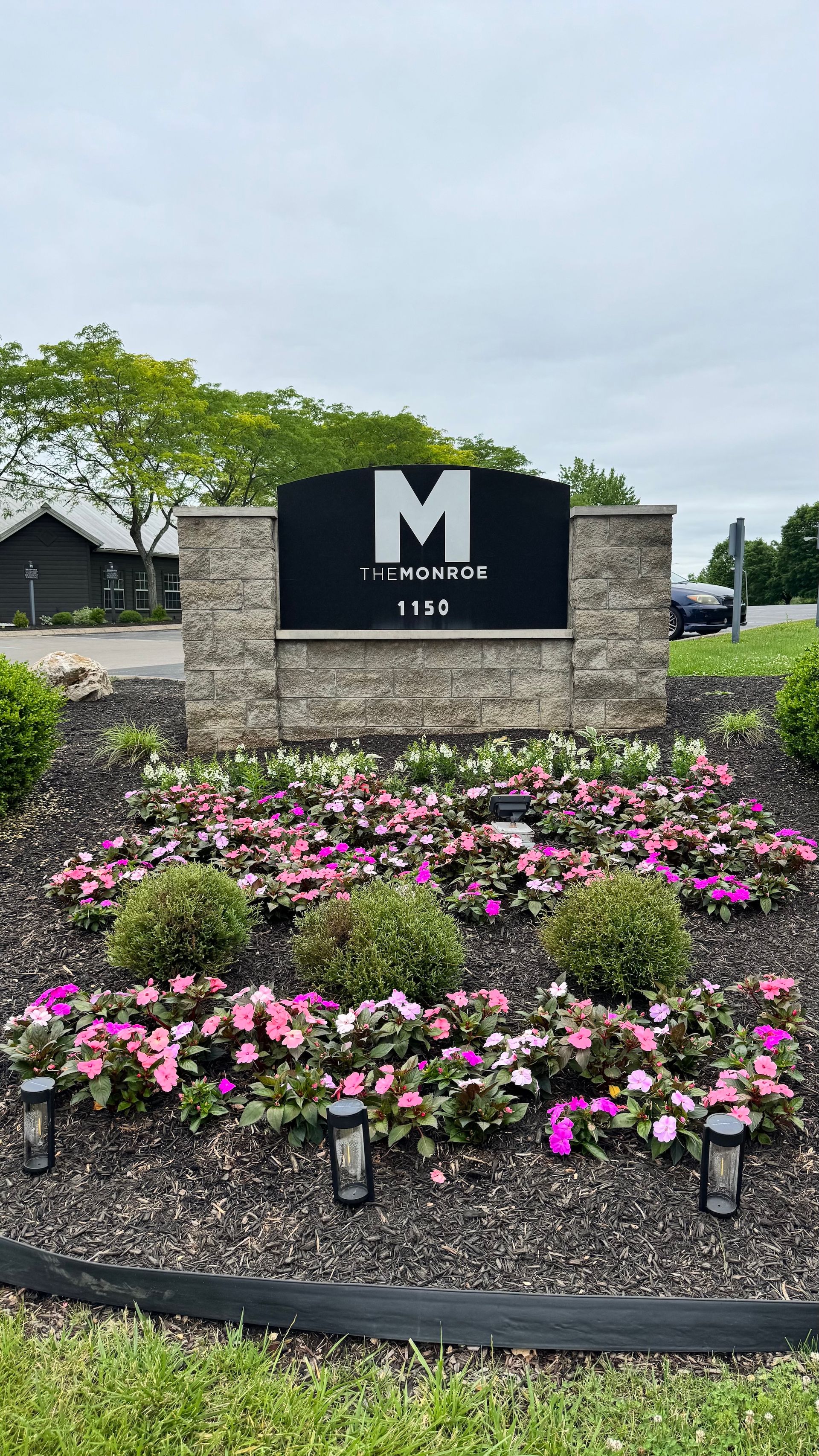 Stone entrance sign with a large white “M,” surrounded by pink flowers and greenery under an overcast sky