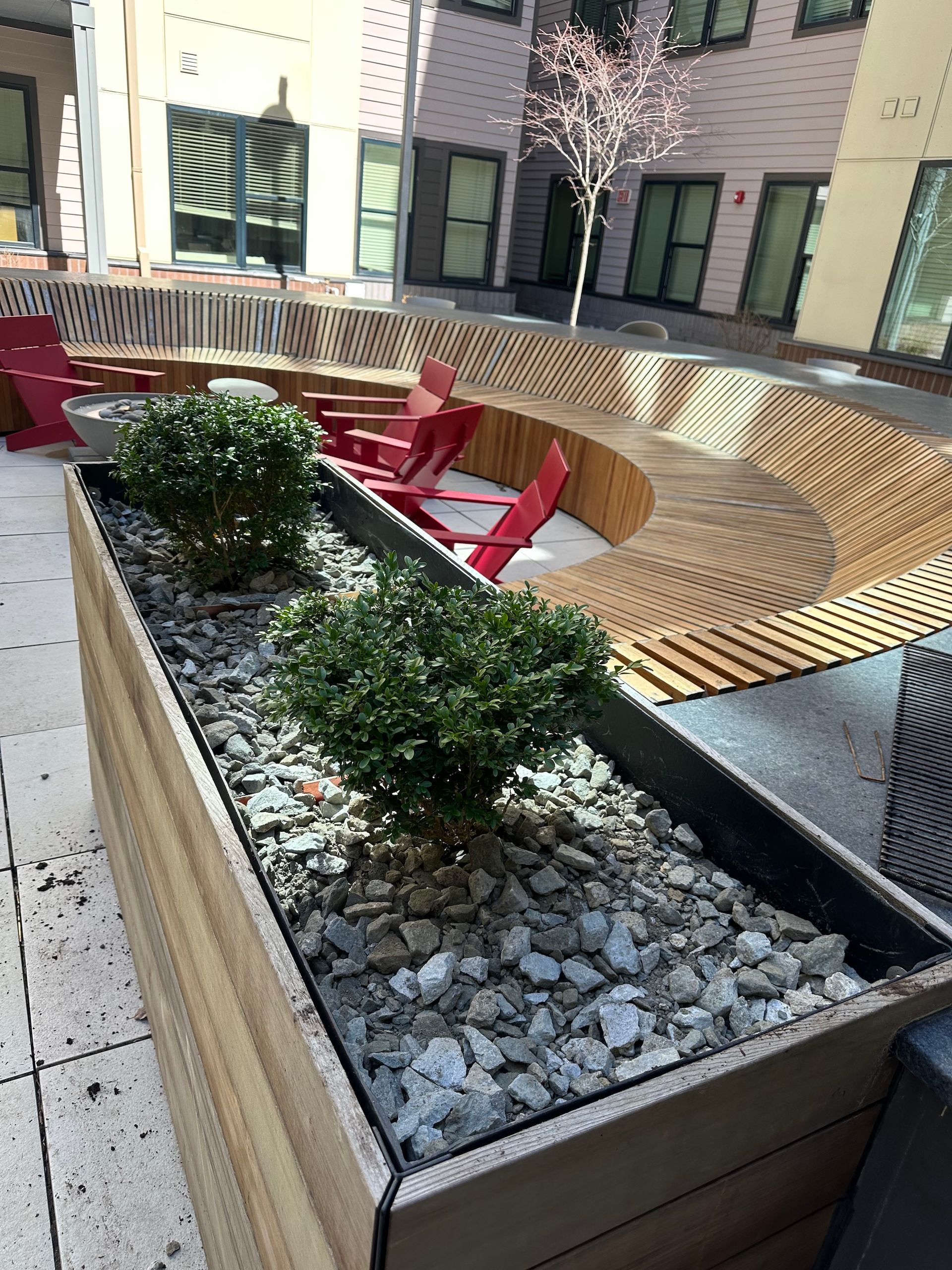 Curved wooden planter with white stones and green shrubs beside red lounge chairs in a courtyard