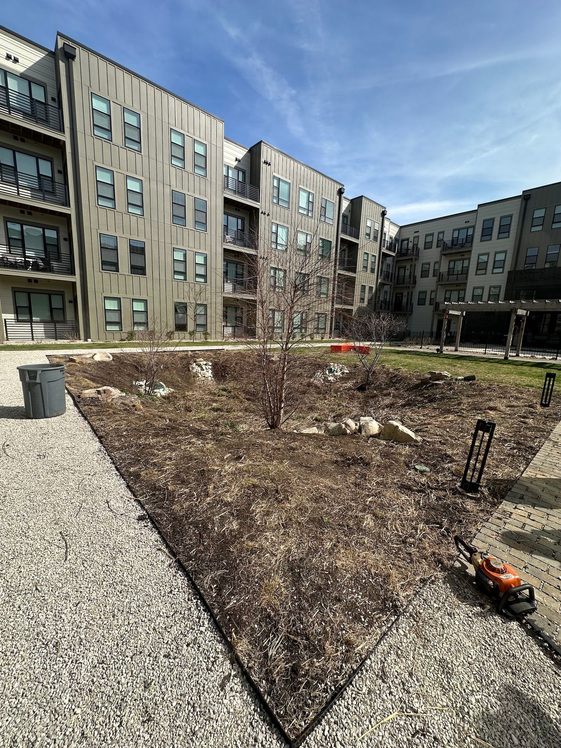 Apartment courtyard with raised gravel planting bed, bare shrubs, and a small orange lawn tool.