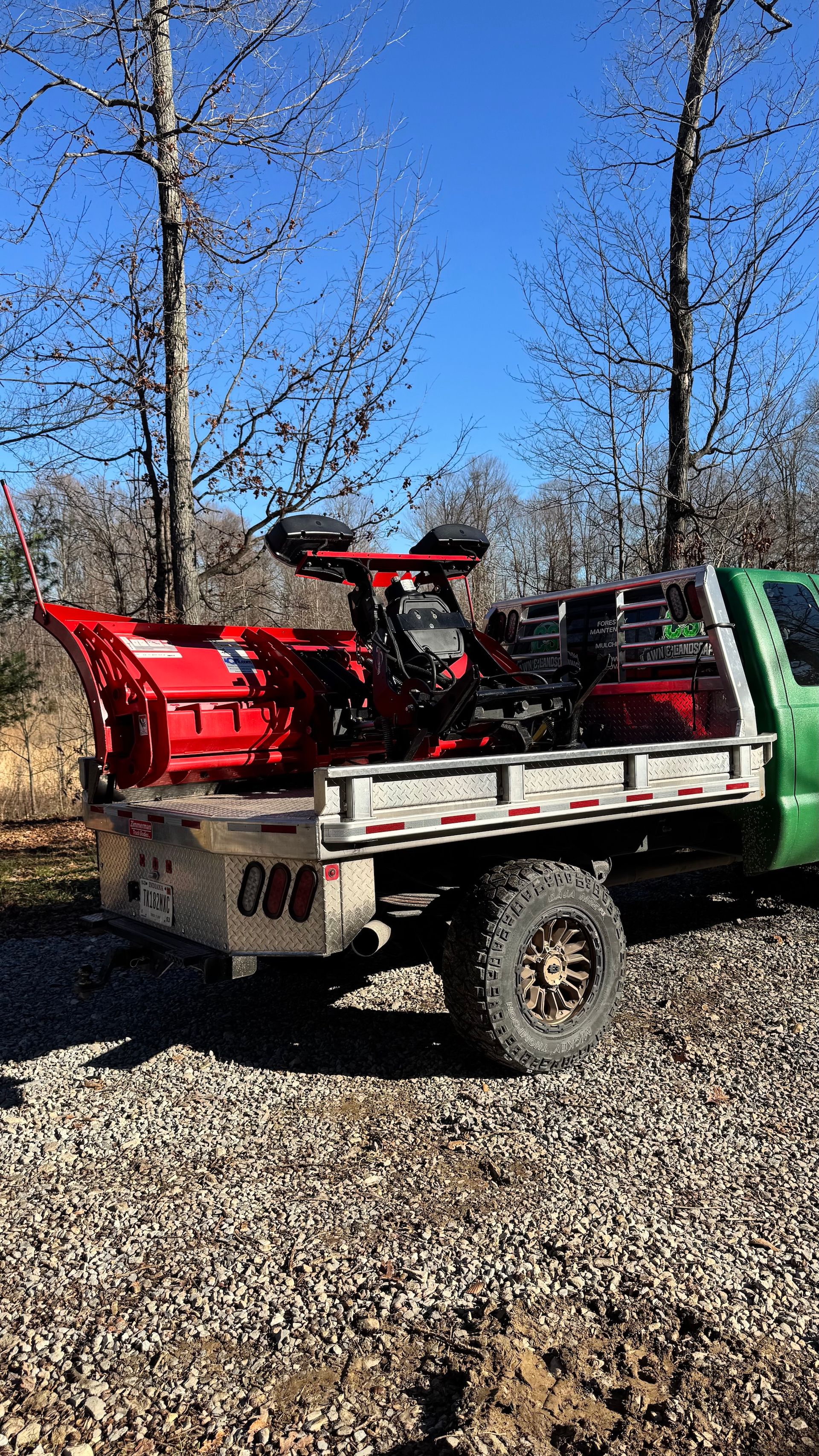 Green utility truck loaded with a red snowblower attachment on a gravel lot under a blue sky