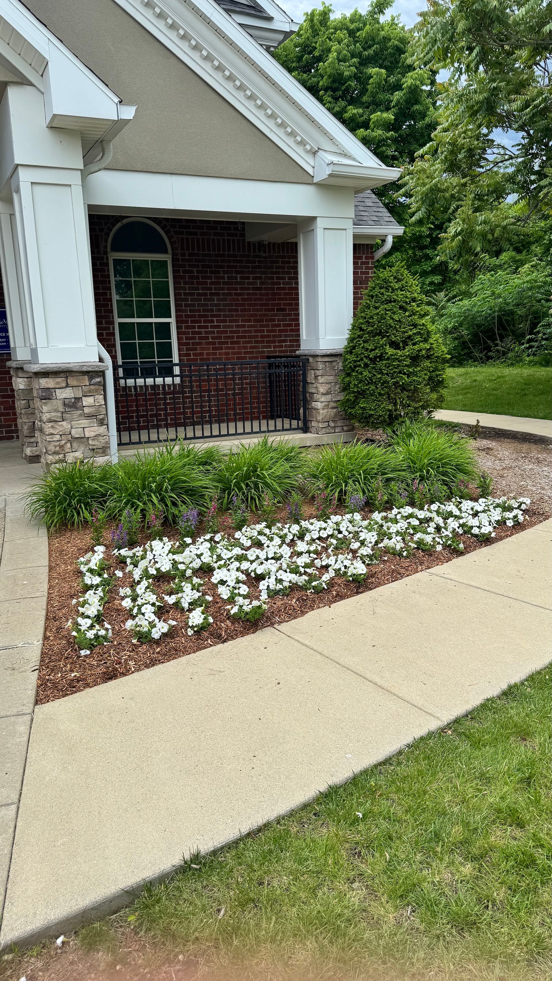 Front porch with black railing, brick steps, and a landscaped flower bed beside a concrete walkway.