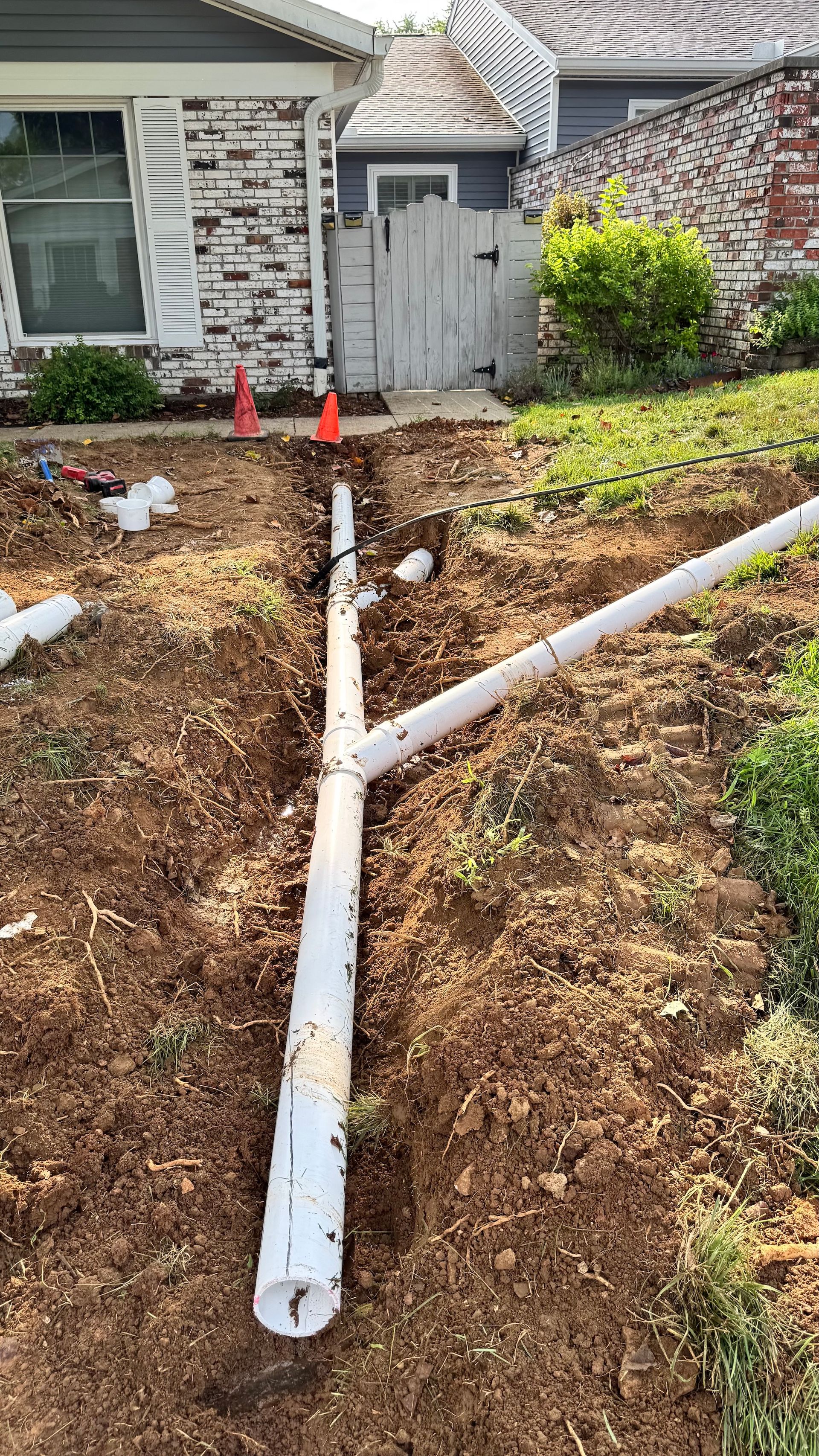 Broken white pipe in a front yard beside a house, with small orange cones near a gate.