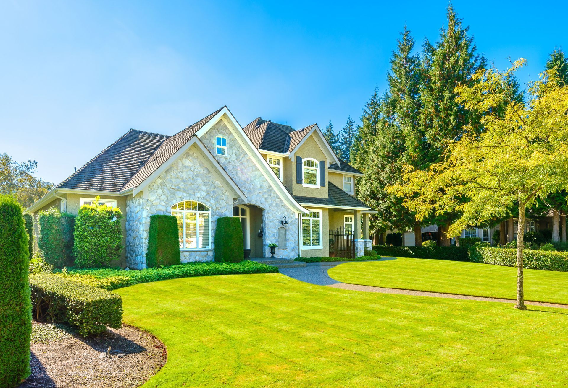 Sunny suburban house with stone façade, green lawn, and tall trees under a clear blue sky