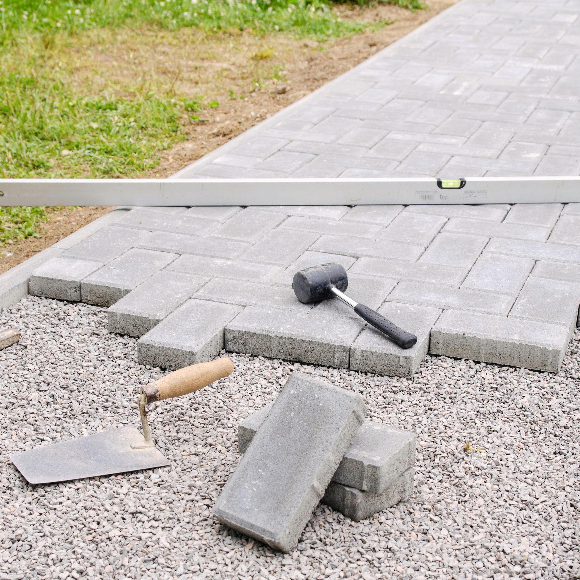 Broken pavers and tools on a walkway under repair, with gravel and grass nearby