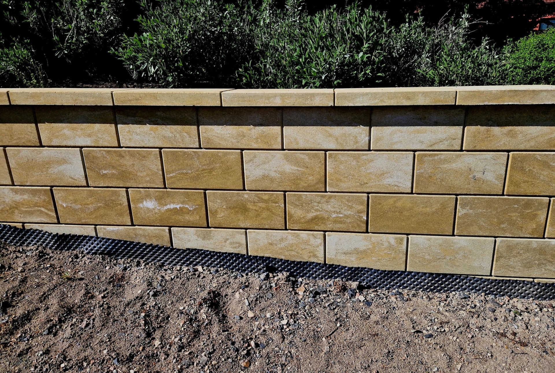 Tan brick retaining wall above a gravel path, with shrubs and grass behind it