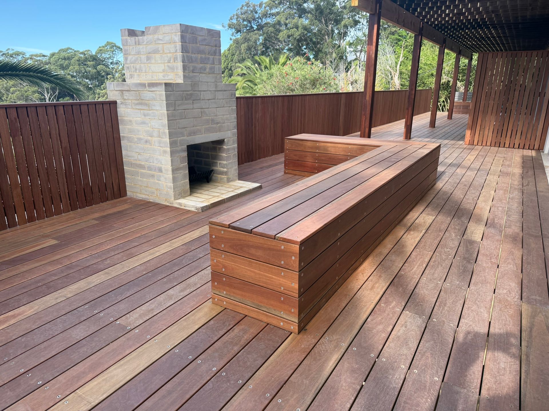 Outdoor wooden deck with built-in bench and stone fireplace, surrounded by a wood privacy fence — Divine Landscape Creations in Berkeley Vale, NSW