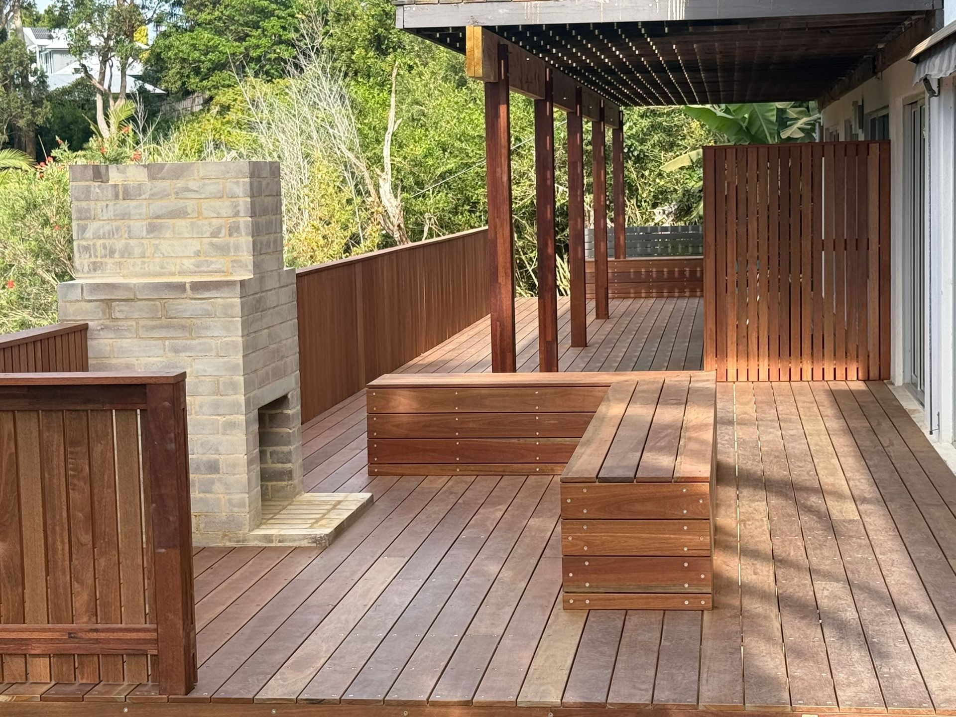 Wooden deck with pergola, built-in bench, and stone outdoor fireplace beside a fence — Divine Landscape Creations in Berkeley Vale, NSW