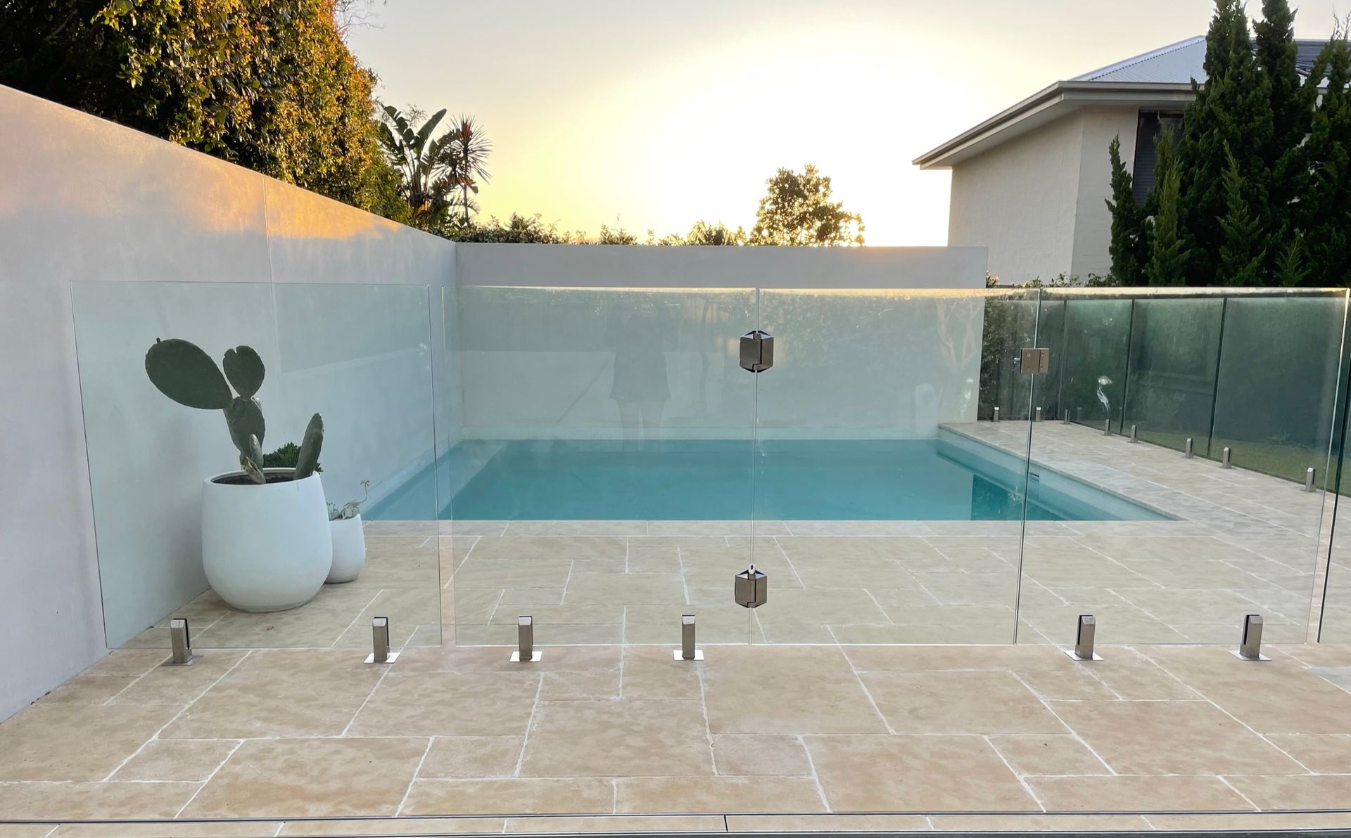 Swimming pool enclosed by glass fencing, beige tiles, white wall, and potted cactus under a sunset.