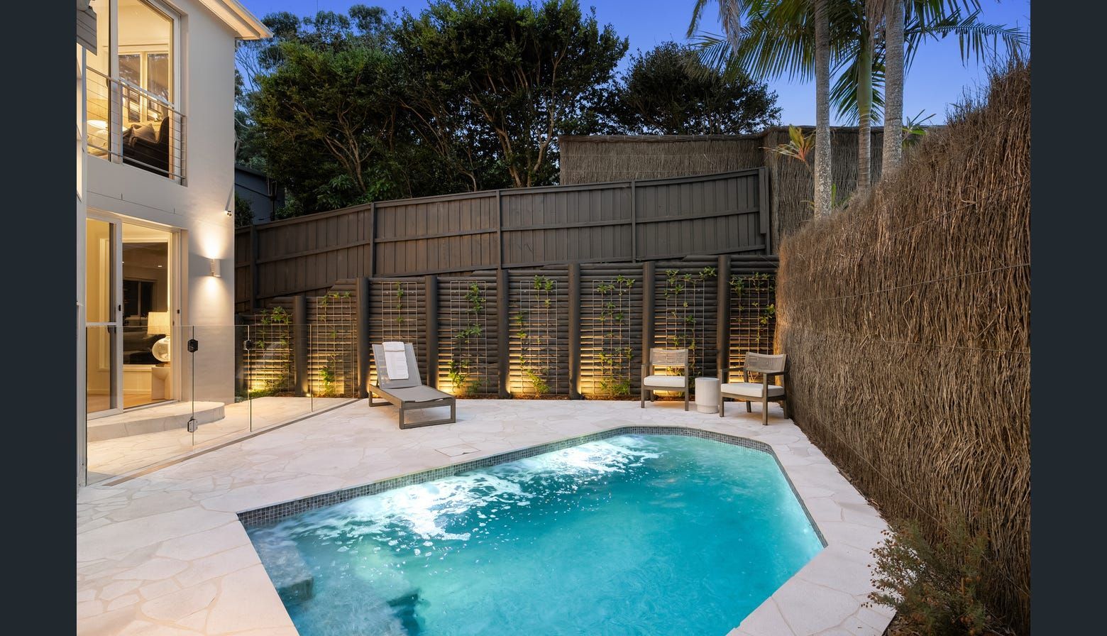 Backyard pool with lounge chairs, surrounded by a fence and lush greenery, evening lighting.