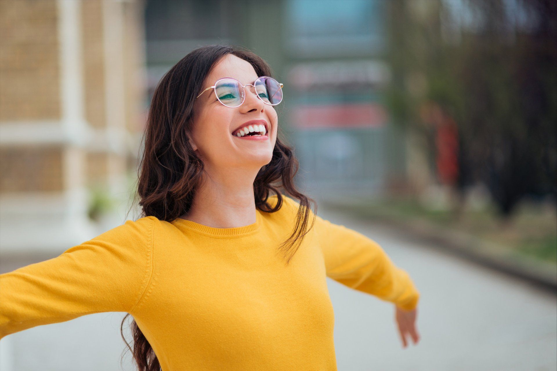 A woman in a yellow sweater and glasses is laughing with her arms outstretched.
