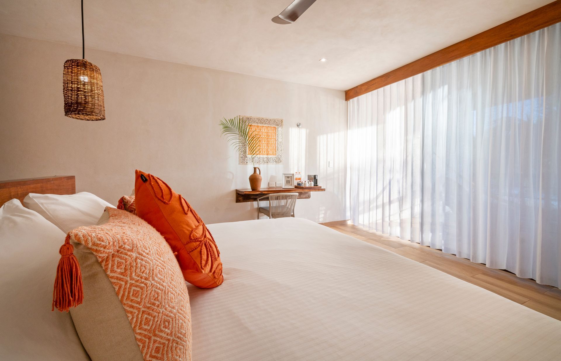 Bedroom with white bedding, orange pillows, sheer curtains, and a wooden table with decor.