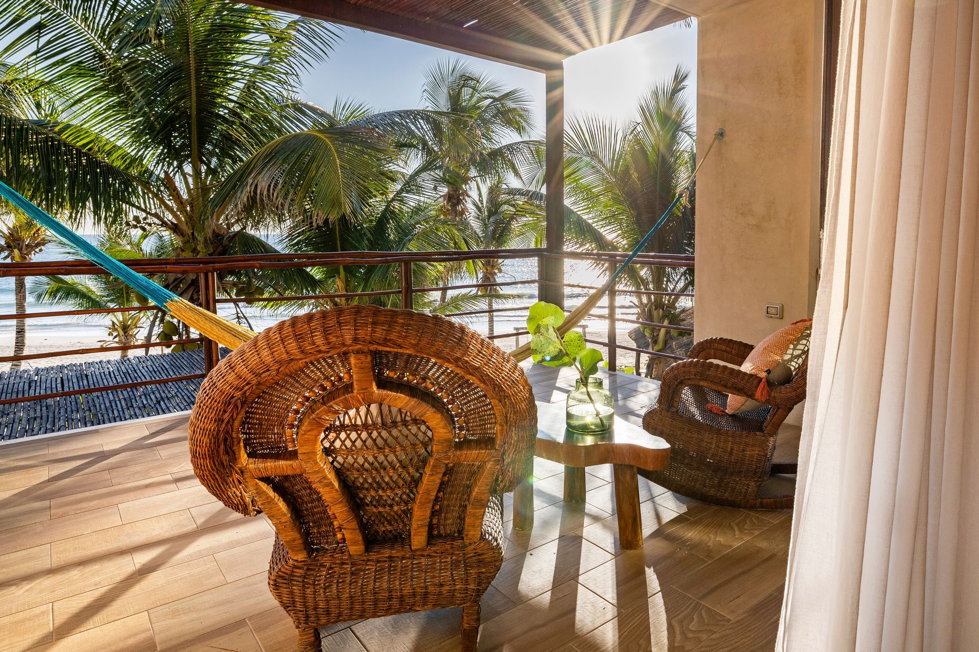Wicker chairs on a balcony overlooking a beach with palm trees and ocean. Bright sunlight.
