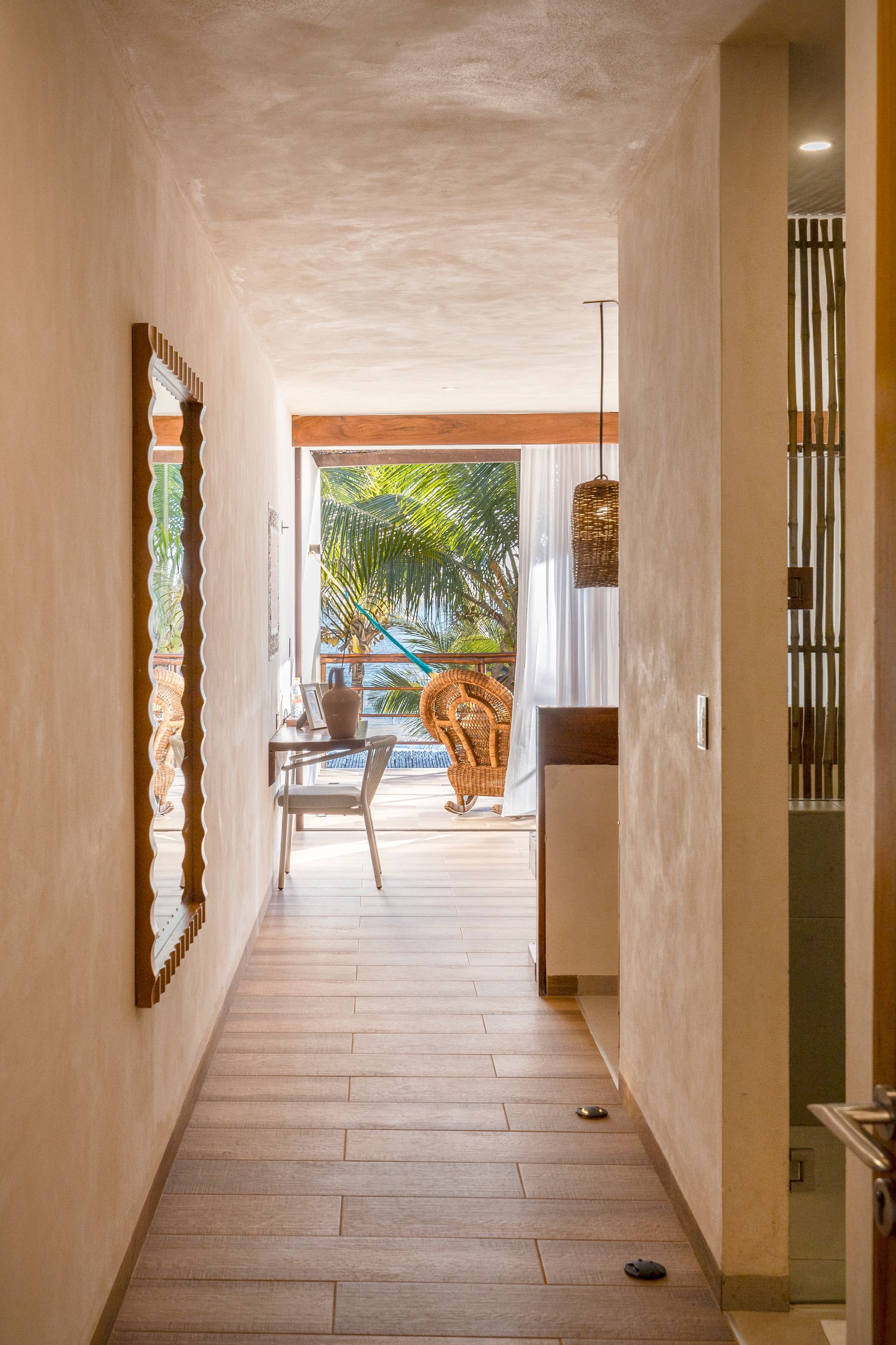 Narrow hallway with wooden floor, mirror on the wall, leading to a bright outdoor view.