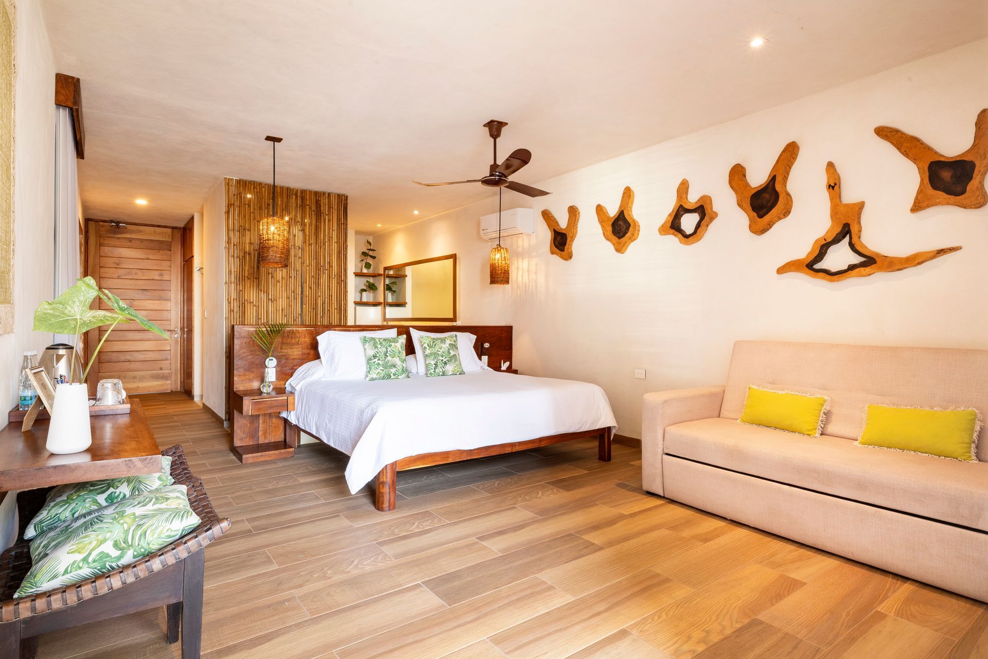Bedroom with a king bed, sofa, and natural wood accents. Features light wood flooring, white walls, and a ceiling fan.