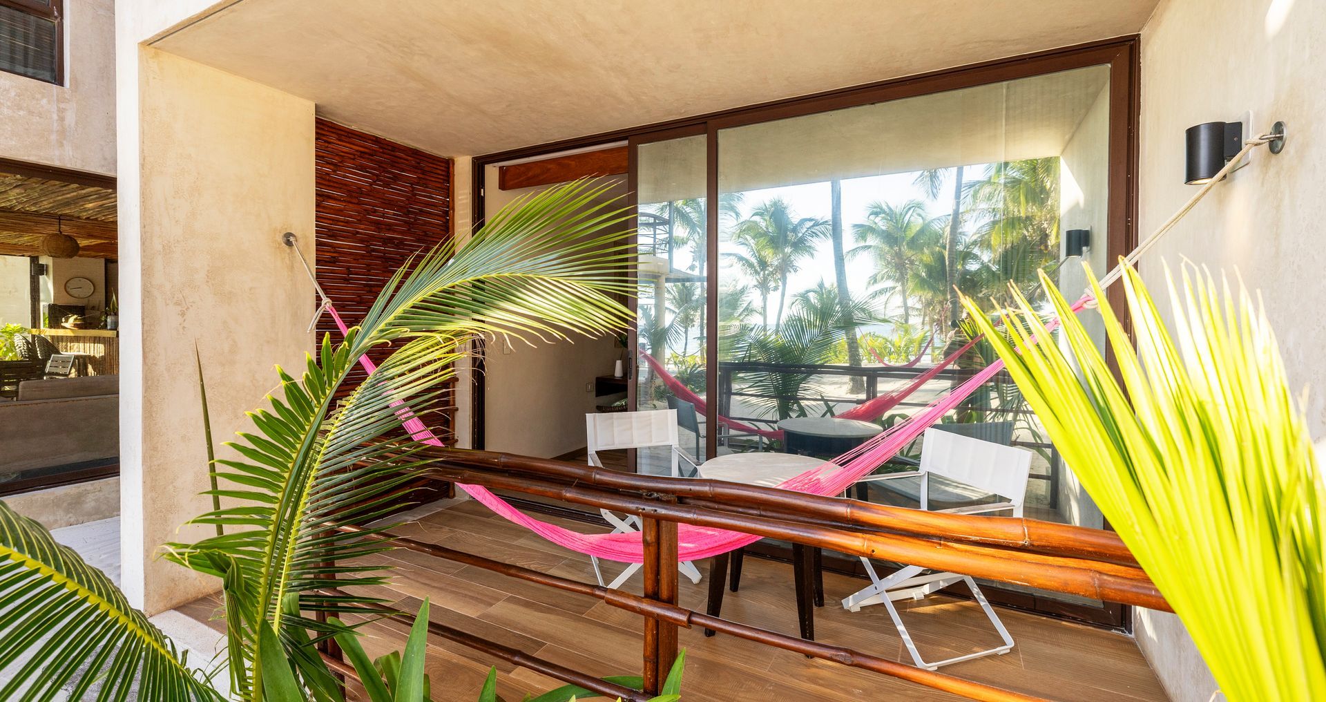 Balcony with hammock, table, chairs, and view of trees and beach. Pink and white decor.