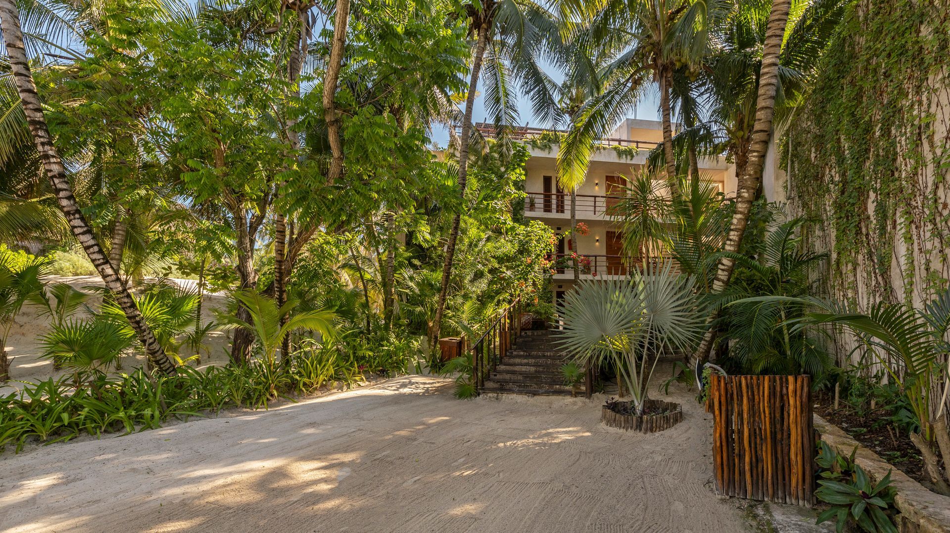 Three-story building with balconies, surrounded by lush tropical vegetation and a gravel driveway.