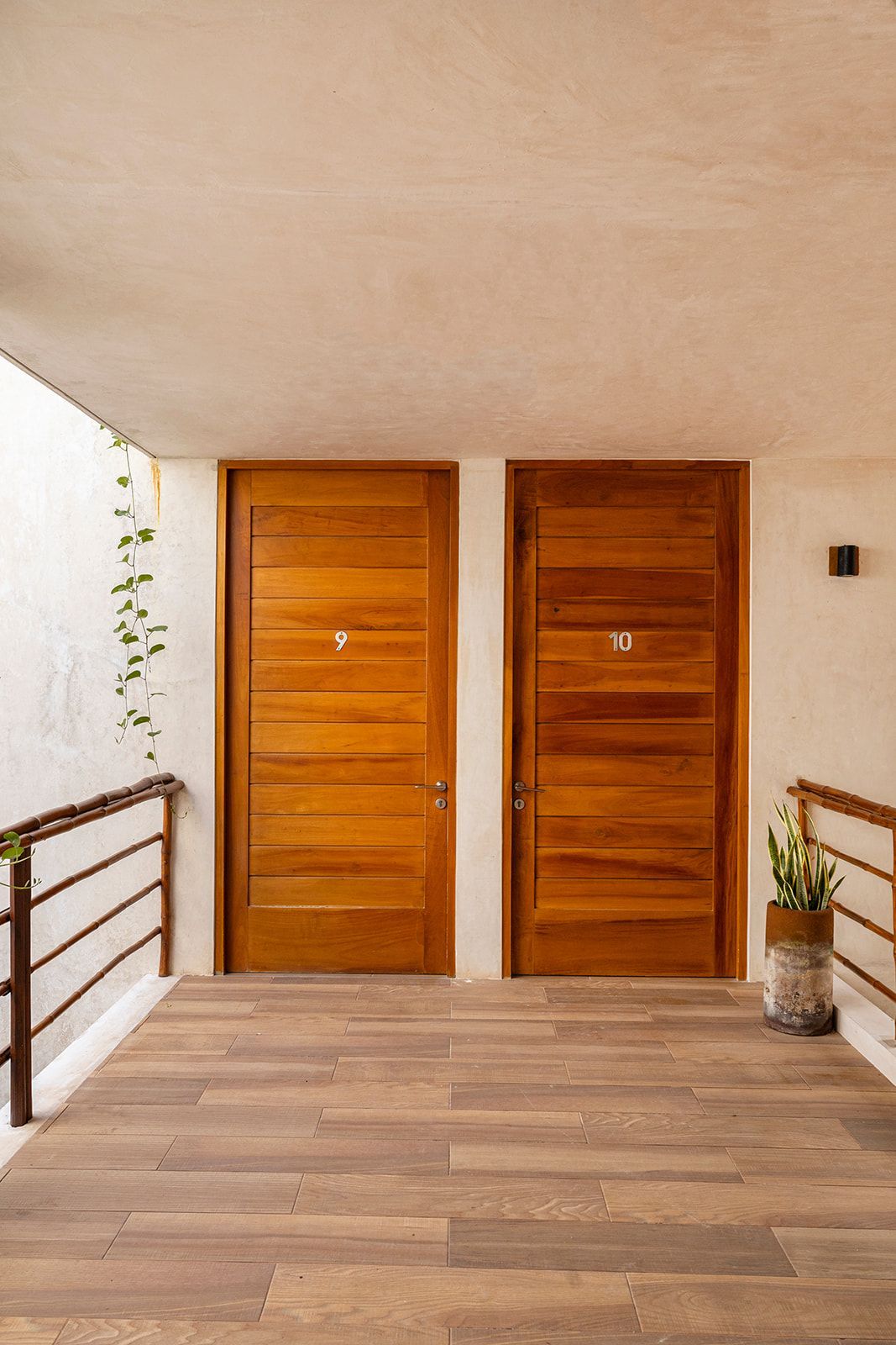 Two wooden doors with numbers 9 and 10 on a walkway with a railing, potted plant, and greenery.