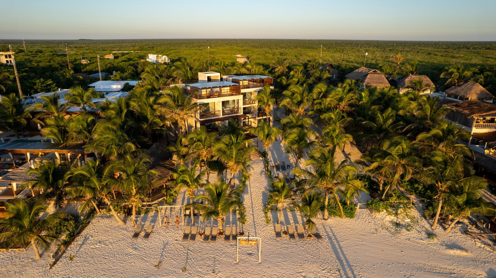 Aerial view of a white sand beach with palm trees leading to a modern building surrounded by lush green forest.