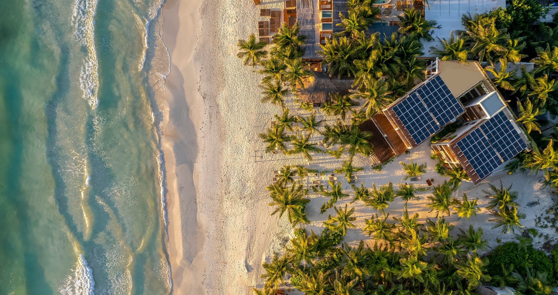 Aerial view: Beach and turquoise water with buildings and solar panels amidst palm trees.