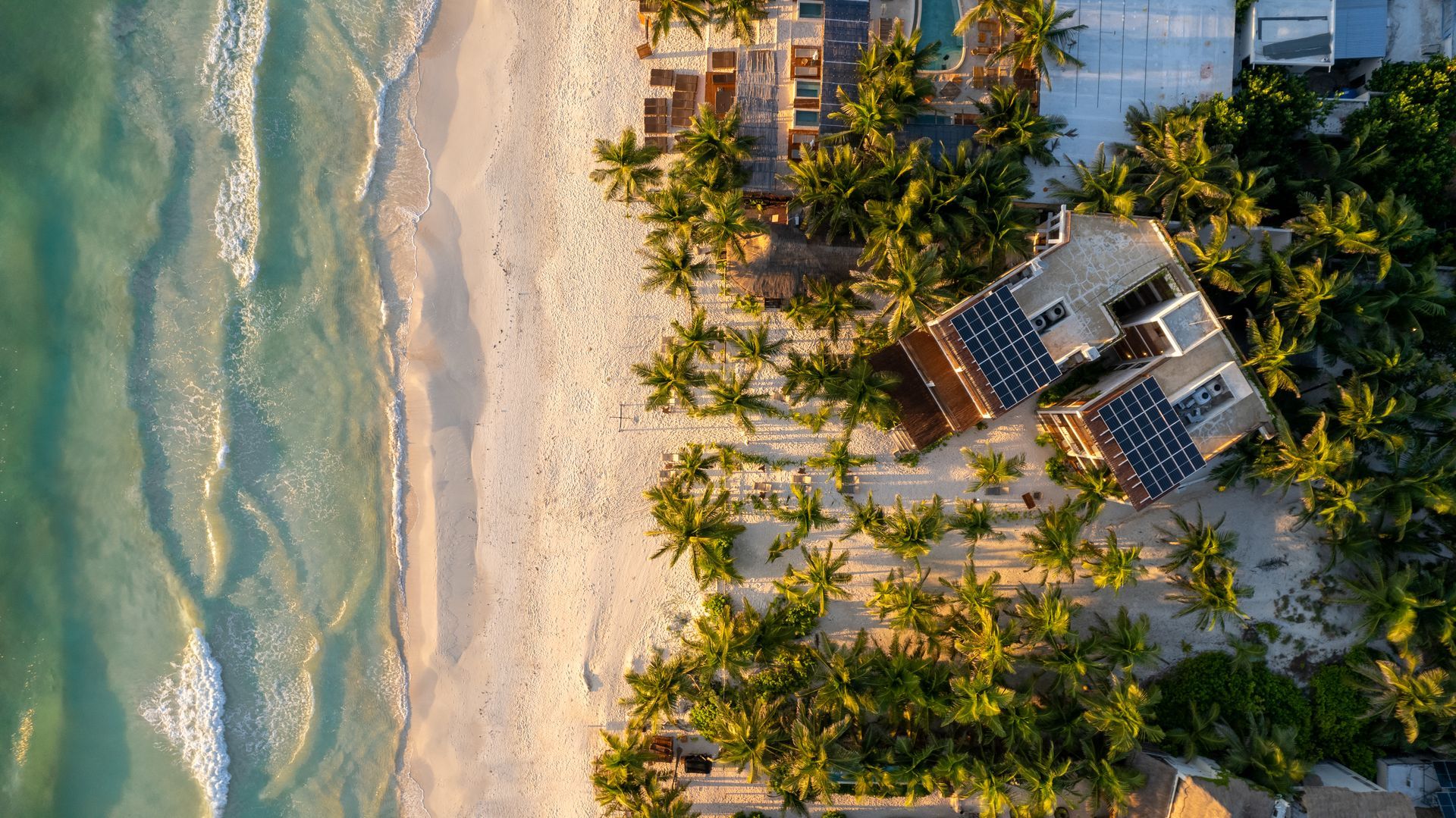 Aerial view of a beach with turquoise water, white sand, and a building with solar panels surrounded by palm trees.