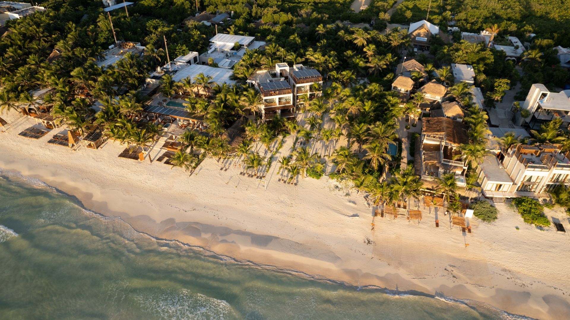 Aerial view of a white sandy beach with turquoise water, lined with palm trees and buildings.