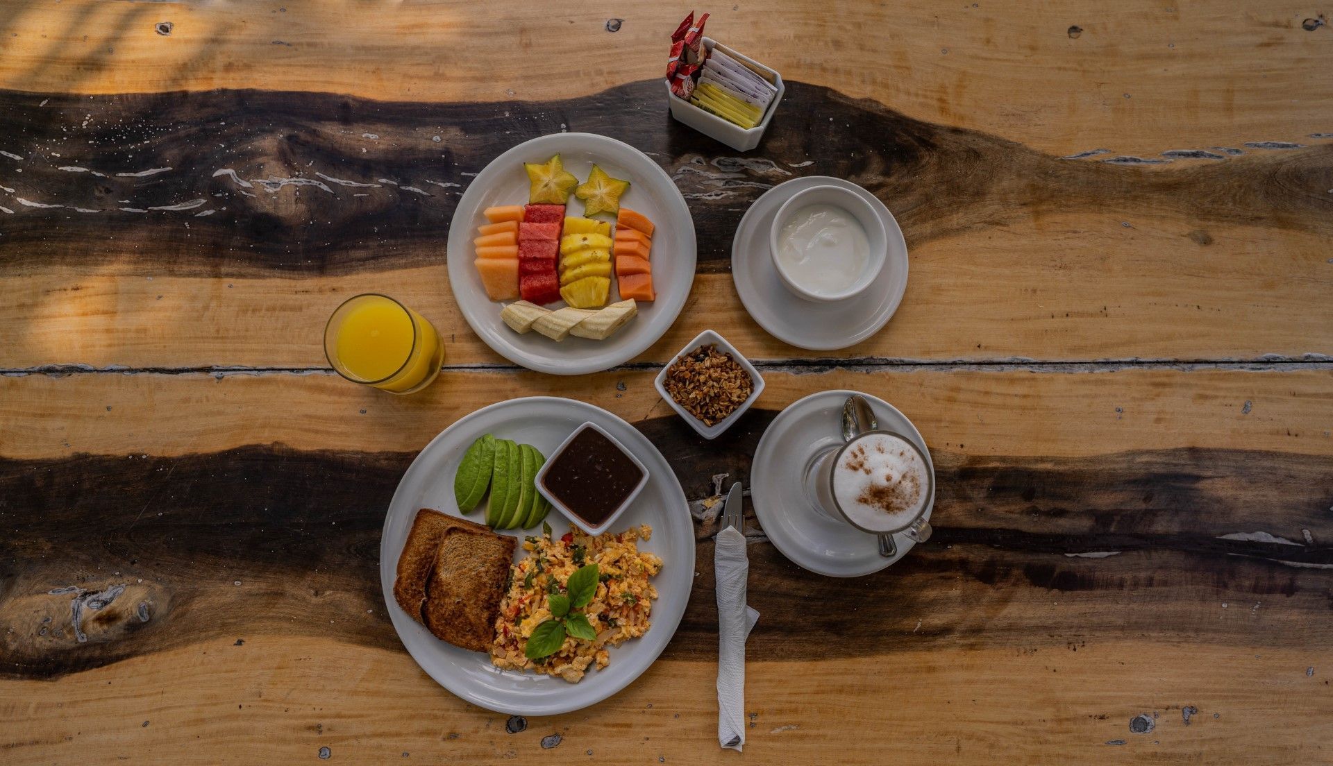 Breakfast spread on a wooden table: fruit plate, scrambled eggs, avocado, toast, coffee, juice, and small containers.