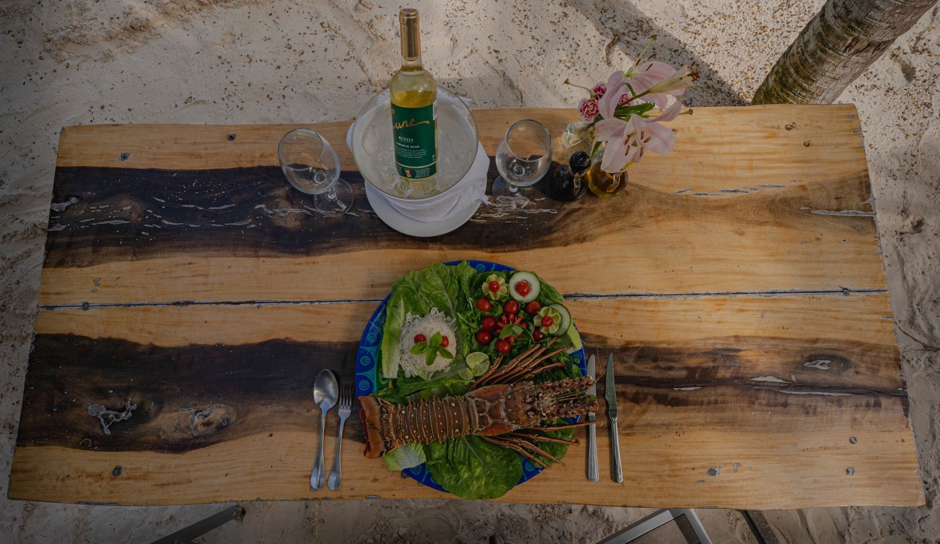 Lobster meal on a wooden table with wine, glasses, and flowers. Outdoors, bright sunlight.