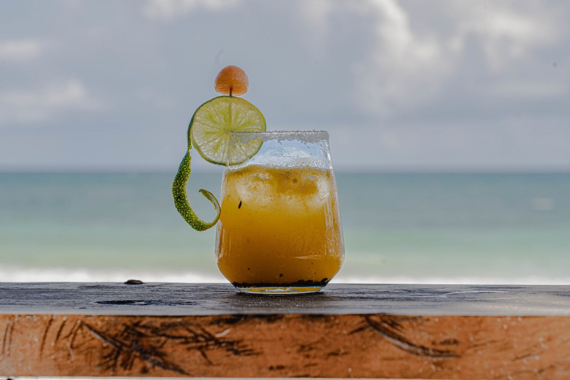 Cocktail with lime and garnish on wooden surface, ocean in the background.