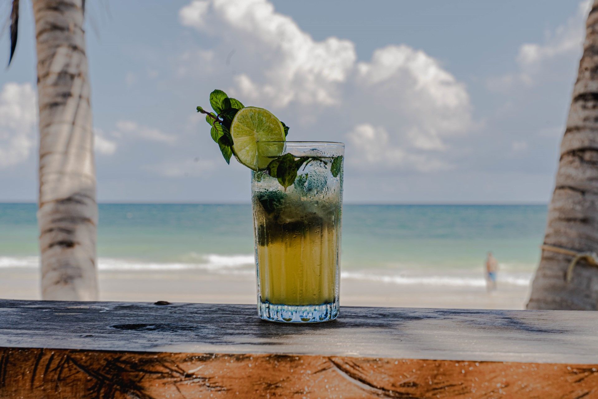 Mojito cocktail on a wooden bar overlooking a beach with palm trees and ocean.